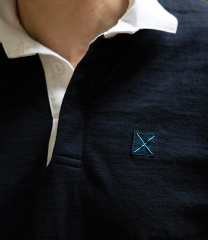 Close-up of a person wearing the McArthur Rugby Jersey by Best Made Co., a navy blue heavyweight cotton piece with a white collar and blue embroidered X on the chest.