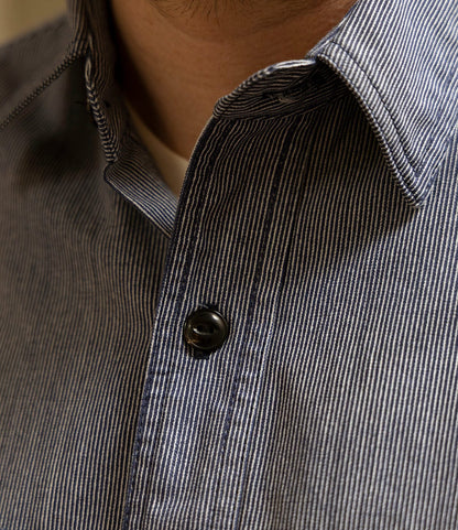 Close-up of a person wearing Best Made Co.'s Indigo Stripe Workshirt, made from Portuguese cotton with triple-needle stitching. Visible are the black button, shirt collar, and part of the lower face and neck.