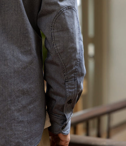 A close-up shows a person's arm in the Best Made Co. Indigo Stripe Workshirt, made from Portuguese cotton with triple-needle stitching, highlighting an oval elbow patch and buttoned cuff beside a blurred indoor wooden railing.