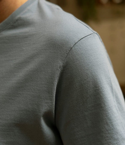 Close-up of a person wearing the Best Made Co. Standard T-Shirt in Overcast Blue, focusing on the shoulder and upper chest. The softly blurred background highlights the fabric and stitching details of this premium cotton tee.