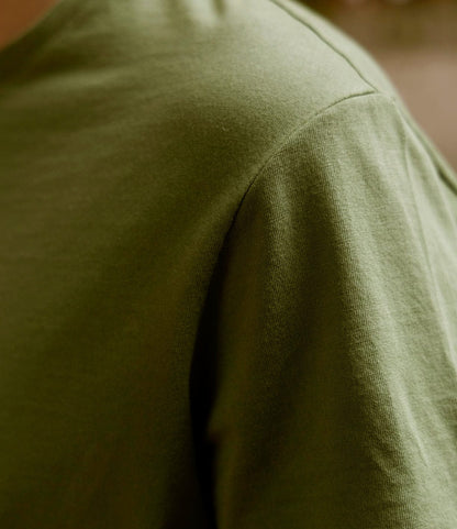 Close-up of a person wearing the Best Made Co. Standard T-Shirt in Olive Drab. The texture of Peruvian tangüis cotton and detailed sleeve stitching are shown, with the shoulder and upper chest in focus against a blurred background.