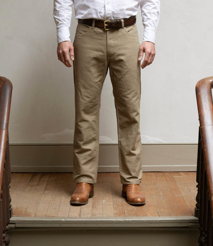 A person wearing Best Made Co.'s Standard 5-Pocket Pant in tan, a white button-up shirt, brown belt, and brown boots stands on a wooden floor between two stair railings, facing forward with hands by their sides.