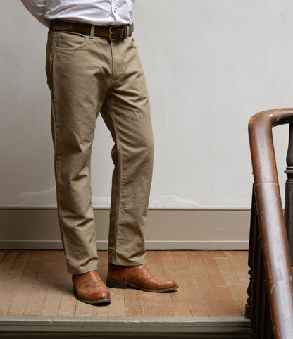 A person stands indoors on a wooden floor, wearing Best Made Co. Standard 5-Pocket Pant in tan, a white dress shirt, a brown belt, and light brown leather boots. Part of a wooden railing appears on the right side.