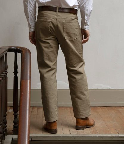 A man stands indoors with his back to the camera, wearing Best Made Co. Standard 5-Pocket Pant in tan, a white shirt, brown belt, and brown shoes beside a wooden staircase on a light-colored wooden floor.