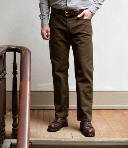 A person stands indoors on a wooden floor wearing Best Made Co.'s Standard 5-Pocket Pant in Deep Olive, paired with a brown belt, striped shirt, and brown leather shoes. Their face is not visible.