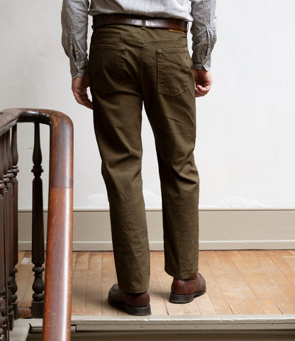 A person stands indoors with their back to the camera, wearing Best Made Co. Standard 5-Pocket Pant in Deep Olive, brown shoes, a striped shirt, and a brown belt, next to a wooden railing and light-colored wall.