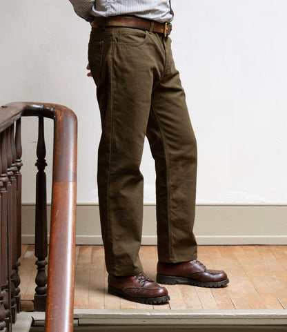 A person wearing Best Made Co.’s Standard 5-Pocket Pant in Deep Olive stands on a wooden floor near a staircase railing. The pants feature a classic straight leg canvas design, paired with a striped shirt and brown leather shoes. Only the lower body is visible.