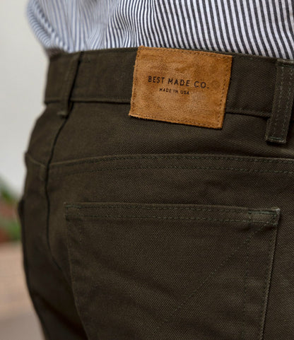 Close-up of a person wearing Best Made Co. Standard 5-Pocket Pant in Deep Olive with a tan leather waistband patch, paired with a white and black striped shirt. Made in USA.