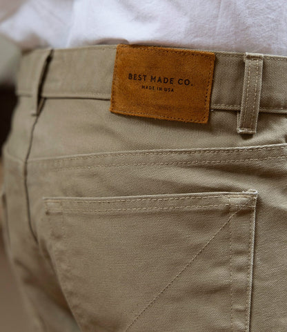 Close-up of the back pocket and waistband of Best Made Co. Standard 5-Pocket Pant Tan, featuring a brown leather patch stamped with the brand name. A white shirt is partially tucked into these straight leg canvas pants.
