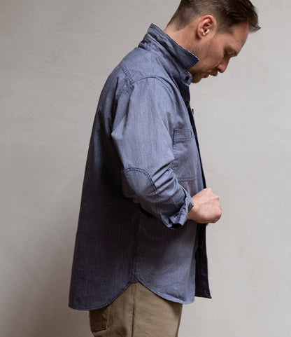 A man in profile, wearing Best Made Co.’s Indigo Stripe Workshirt made from Portuguese cotton with triple-needle stitching, pairs it with beige pants as he looks down and adjusts a shirt button against a plain background.