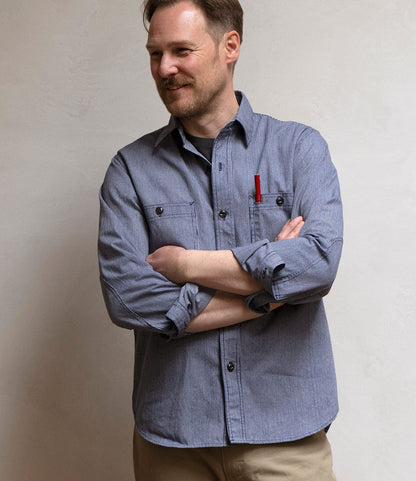 A man with light brown hair and a beard stands smiling with arms crossed, wearing Best Made Co.’s Indigo Stripe Workshirt (Portuguese cotton, red pen in chest pocket) and beige pants against a plain light-colored wall.