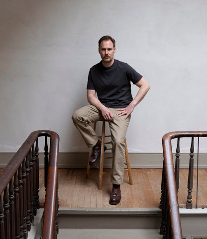 A man in a black t-shirt and Best Made Co. Standard 5-Pocket Pant Tan sits on a wooden stool at the end of a hallway with wooden floors and railings, against a plain, light-colored wall.