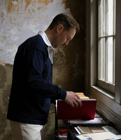 Wearing the Best Made Co. McArthur Rugby Jersey, a man stands by a window in a room with peeling wallpaper, gazing down at a large red book on a paper-filled table as soft daylight fills the space.