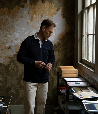 Wearing a McArthur Rugby Jersey by Best Made Co., a man stands by a window in a dim room with peeling wallpaper, holding a pen and looking down at a small wooden box on a metal shelf filled with papers and folders.