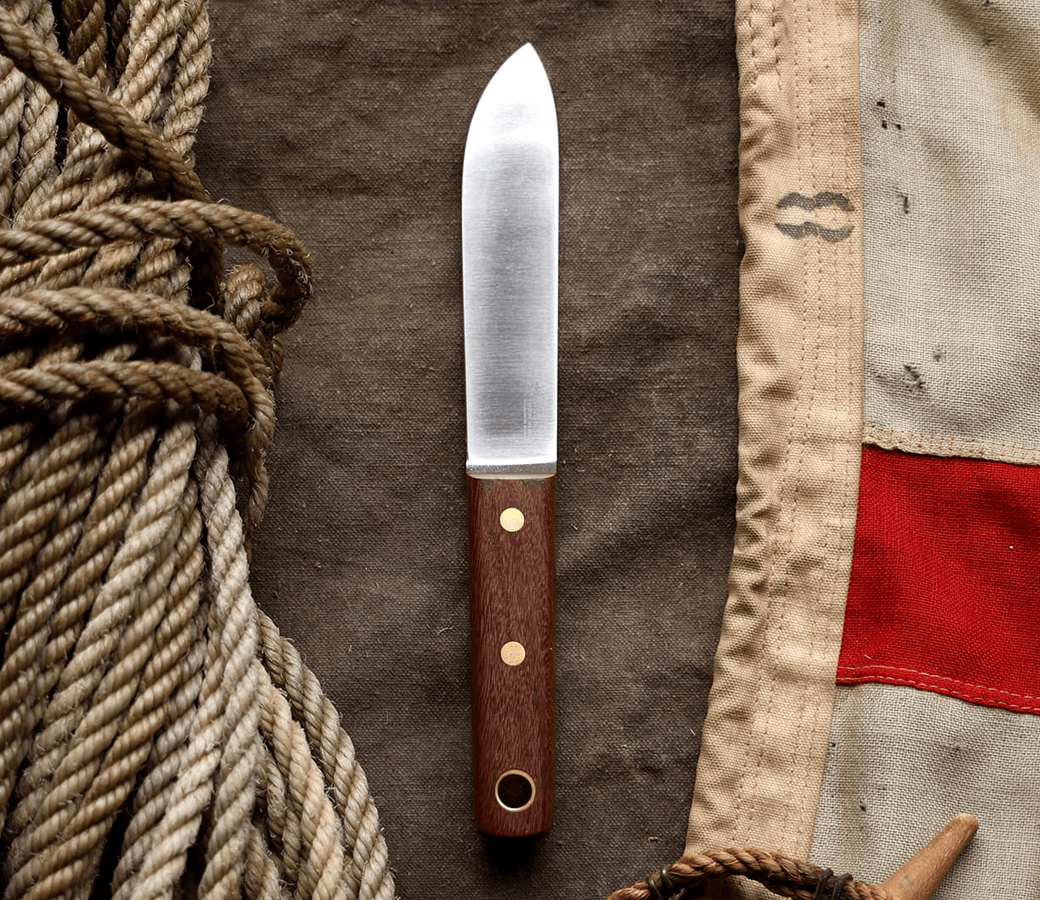 A fixed-blade knife with a wooden handle lies on brown fabric between coiled rope and a beige canvas with a red stripe.