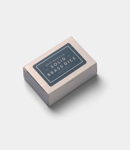 A small rectangular box with a blue Best Made Co. label reading Solid Brass Dice rests on a light gray surface, its minimalist design suggesting the premium quality of the machined brass dice within.