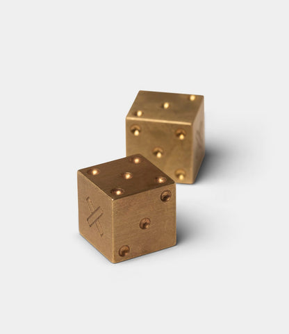 Two Solid Brass Dice from Best Made Co. rest on a white background. The front die shows three dots and an engraved X-like symbol, while the rear, slightly blurred die features five dots.