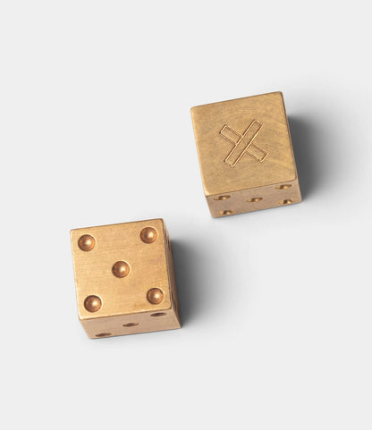 Two Best Made Co. Solid Brass Dice rest on a light gray surface—one shows four pips, while the other features a machined brass face with an engraved X in place of standard dots.