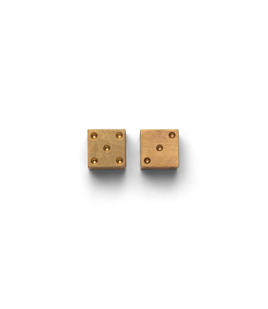 Two Best Made Co. Solid Brass Dice rest on a light gray surface—one shows four pips, while the other features a machined brass face with an engraved X in place of standard dots.