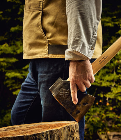 A person stands outdoors by a tree stump and holds the Best Made Co. Old Gold No. 1 hand-forged axe, surrounded by green foliage.
