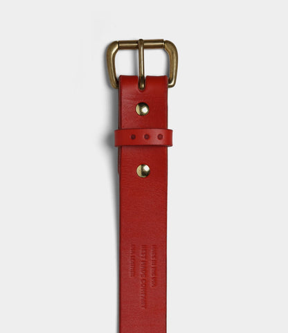 The Famous Red Belt by Best Made Co. is a bridle leather belt in red with a brass buckle, two gold rivets, and two adjustment holes. Embossed text appears near the end; shown vertically on a white background. Made in USA.