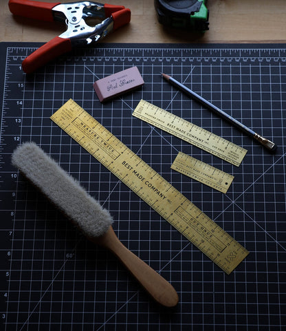A Best Made Co. Measure Twice 6 inch Solid Brass Ruler, plus a brush, two other brass rulers, pencil, pink eraser, hole punch, and green tape measure are arranged neatly on a black cutting mat in soft light.
