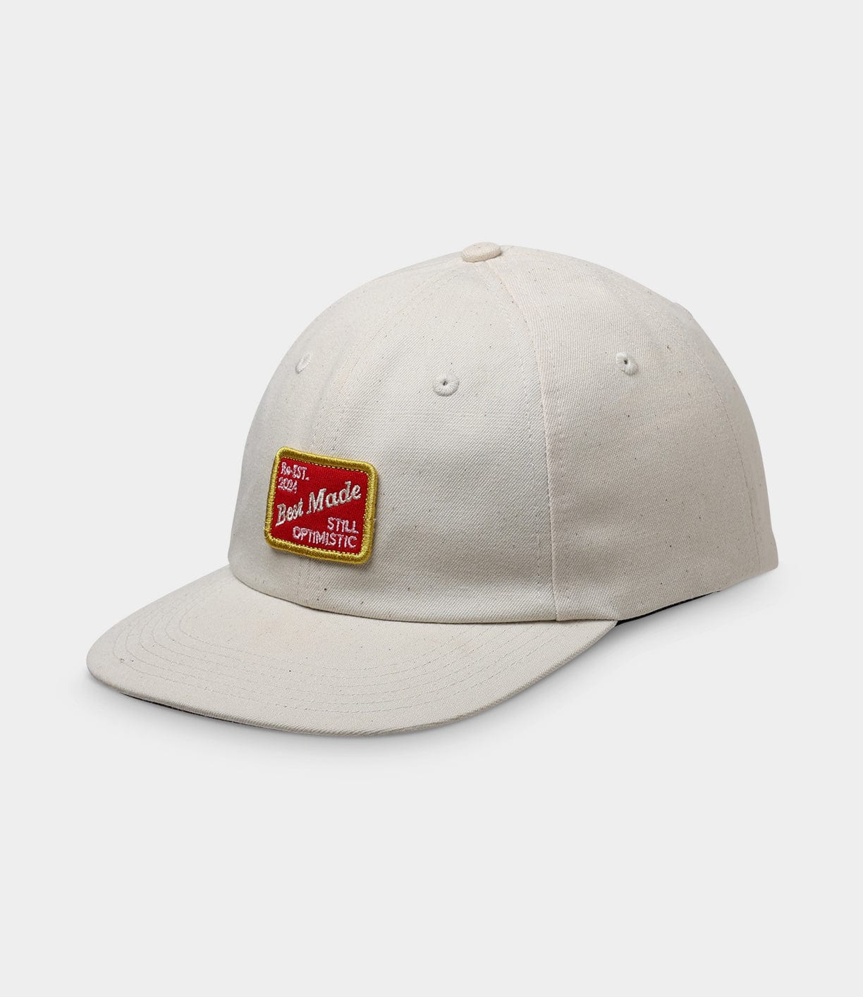 The Selvedge Natural Denim Ball Cap by Best Made Co. is cream-colored with a curved brim, a red and yellow "Best Mode Still Optimistic" patch, and a leather adjustment strap at the back.