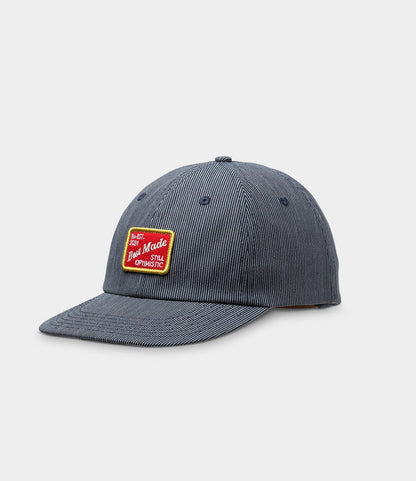 The Best Made Co. Small Stripe Indigo Ball Cap features blue and white hickory stripe denim, a curved brim, and a front red-and-yellow "Good Made, Still Optimistic" patch, shown on a plain white background.