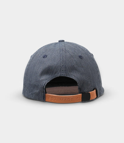The Best Made Co. Small Stripe Indigo Ball Cap is seen from the back, showcasing blue and white hickory stripe denim and a brown leather strap with embossed text for size adjustment.