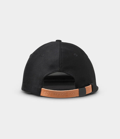 The Military Cord Ball Cap Black by Best Made Co. is shown from the back, highlighting a leather adjustment strap and tan leather patch embossed with the brand logo, set against a plain light gray background.