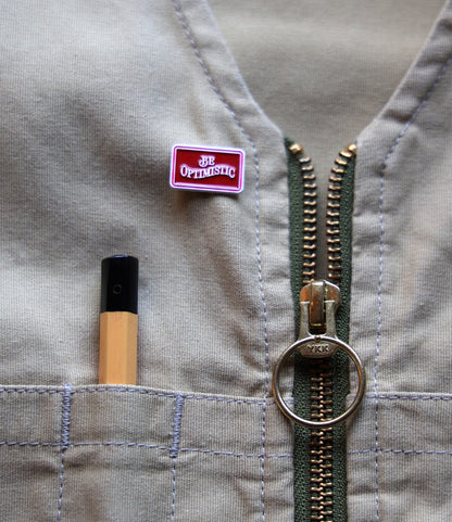 A beige fabric with a zipper, a pen in a stitched pocket, and Best Made Co.'s Be Optimistic Pin—a small red enamel pin that reads “BE OPTIMISTIC.”.