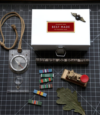 A Best Made Co. Type 6 Strongbox (Re-Founder's Edition), a compass on a lanyard, military ribbons, a brass whistle with red button, winged badge, and oak leaf sit on a black grid cutting mat—perfect for workshop organization.