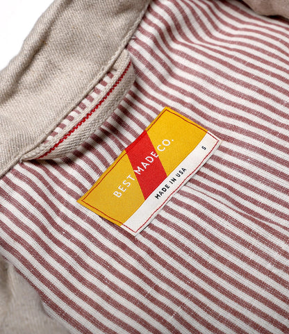 Close-up of the Selvedge Natural Studio Jacket collar with red and white vertical stripes, highlighting American craftsmanship. A yellow and red Best Made Co. label reads MADE IN USA, with size S indicated on the right.