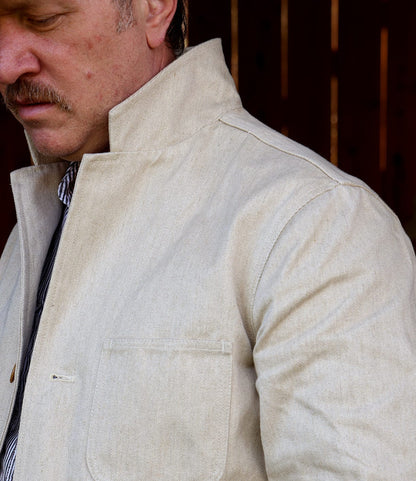A man with a mustache wears the Selvedge Natural Studio Jacket by Best Made Co., made from organic cotton-hemp, featuring a high collar and front pocket. Only his lower face and upper body are visible against a dark background.