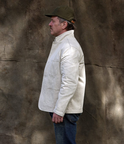 A man stands in profile against a textured brown background, wearing the Selvedge Natural Studio Jacket by Best Made Co., blue jeans, and an olive green baseball cap—highlighting American craftsmanship.