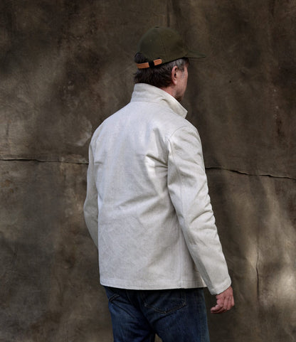 A person wearing the Selvedge Natural Studio Jacket by Best Made Co., blue jeans, and an olive green cap stands with their back to the camera before a textured brown backdrop.