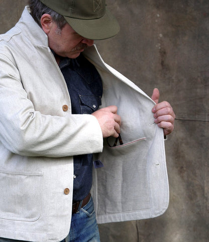 Wearing a Best Made Co. Selvedge Natural Studio Jacket and a green cap, a person opens the jacket to access an inner pocket, holding glasses against a plain, neutral background.