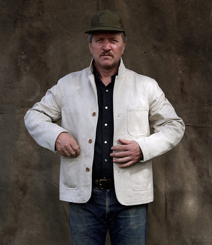 A man in a green cap, dark shirt, and blue jeans stands against a brown backdrop, adjusting his Selvedge Natural Studio Jacket by Best Made Co. and looking at the camera with a neutral expression.