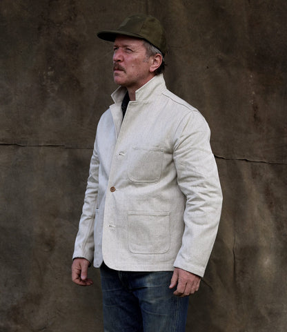 A man with a mustache wears the Best Made Co. Selvedge Natural Studio Jacket, a green cap, and jeans, standing before a textured brown wall and gazing to the side with a serious expression that reflects American craftsmanship.