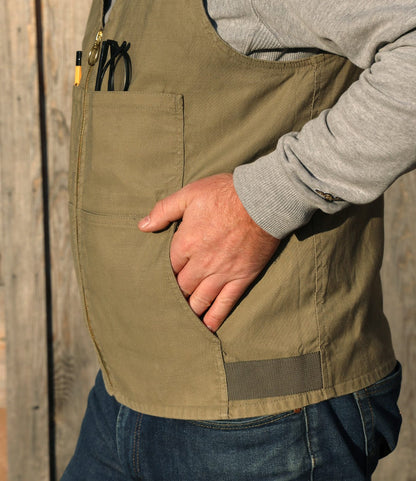 A person stands sideways with one hand in their PB-S’s Utility Vest Black by Best Made Co., layered over a gray long-sleeve shirt and blue jeans. The background features wooden planks.