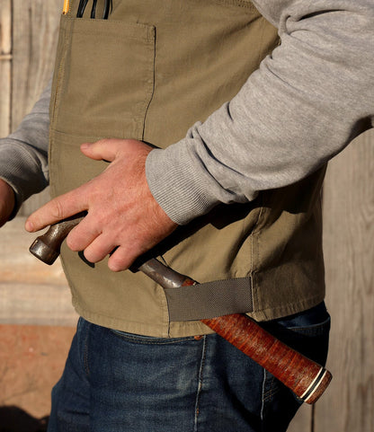 Wearing Best Made Co.’s PB-S’s Utility Vest Black over a gray long-sleeve shirt, a person tucks a brown-handled hammer into their vest, with a wooden backdrop that hints at an outdoor or workshop setting.