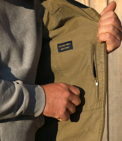 A person wearing a grey sweatshirt opens a black PB-S’s Utility Vest by Best Made Co. to reveal an inside pocket with a zipper and a label that reads ALL GOOD THINGS MUST START.