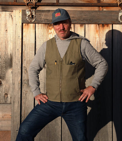 A man stands confidently in sunlight before a rustic wooden wall, wearing blue jeans, a gray hoodie, and the PB-S’s Utility Vest Black by Best Made Co., crafted from durable Japanese cord fabric.