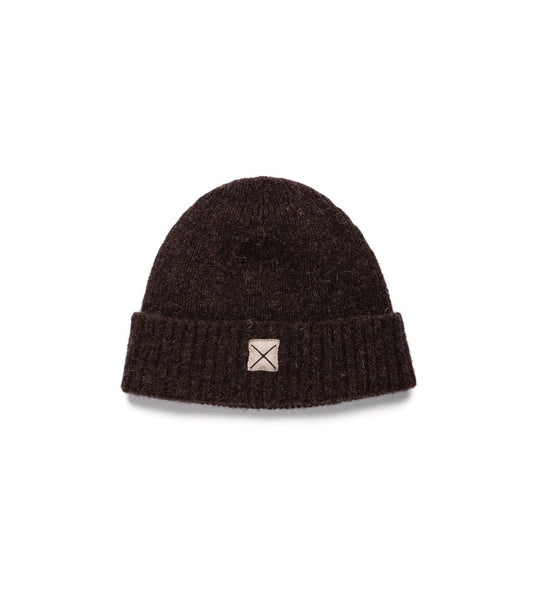 The Best Made Co Shetland Cap of Courage is a dark brown undyed wool cap with a folded brim and a small beige square patch featuring an X on the front, shown on a plain white background.