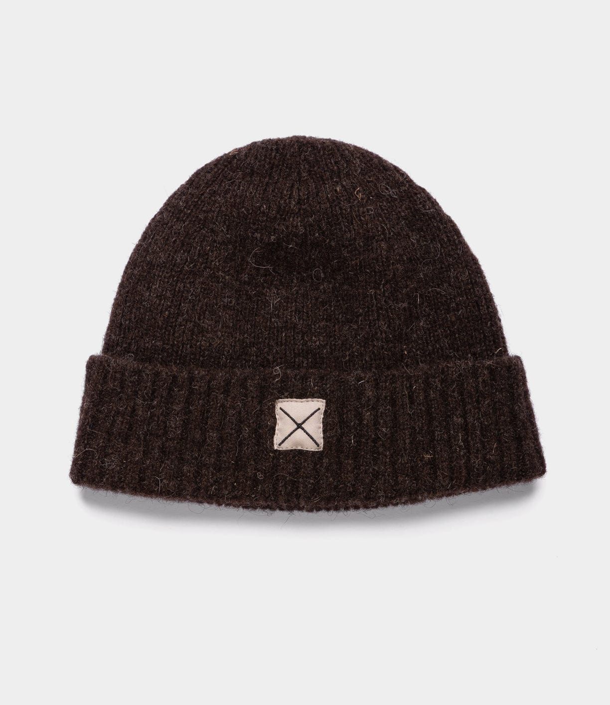 The Best Made Co Shetland Cap of Courage is a dark brown undyed wool cap with a folded brim and a small beige square patch featuring an X on the front, shown on a plain white background.