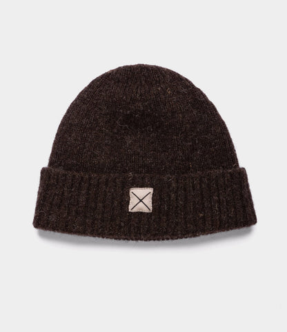 The Shetland Cap of Courage by Best Made Co. is a dark brown weather-resistant beanie with a wide folded brim and a beige patch with a stitched X on the front, offering classic style and cozy warmth. Displayed on a plain light background.