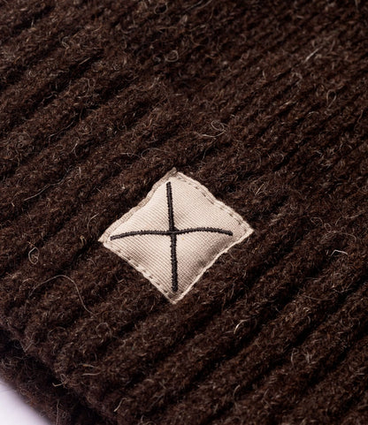 Close-up of the Shetland Cap of Courage by Best Made Co., featuring undyed brown knitted wool and a beige diamond patch with a black stitched cross at the center.