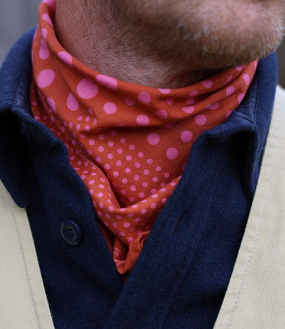A close-up of a person’s neck wearing the Best Made Co. Trademark Bandana Red, styled as a scarf with pink polka dots, paired with a navy blue jacket and cream-colored strap. The lower face and short beard are also visible.