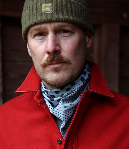 A man with a reddish-blonde mustache and beard wears an olive green beanie, red wool jacket, and the Best Made Co. Trademark Bandana Blue around his neck as a scarf. He faces the camera with a neutral expression.