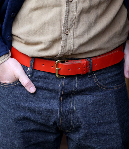 Wearing a tan button-up shirt, dark blue jeans, and the Famous Red Belt by Best Made Co.—a striking bridle leather belt made in the USA with a brass buckle. Only their torso and upper legs are visible, hands tucked in pockets.